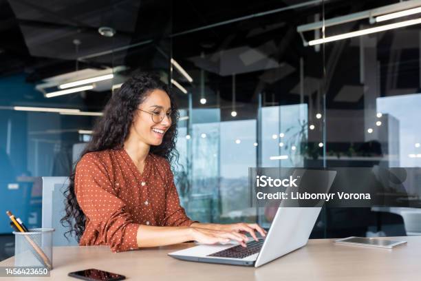 Happy And Smiling Hispanic Businesswoman Typing On Laptop Office Worker With Curly Hair And Glasses Happy With Achievement Results At Work Inside Office Building Stock Photo - Download Image Now