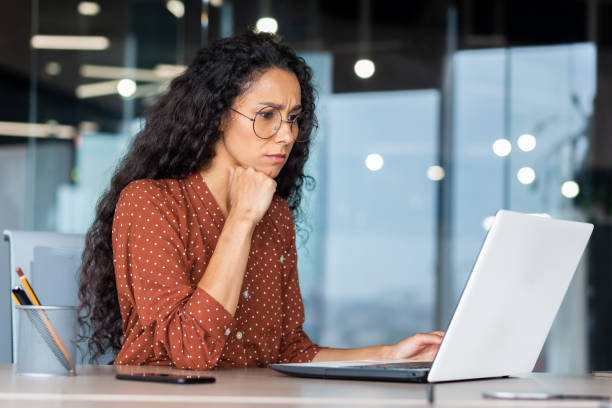 istock Serious thinking Hispanic businesswoman working inside modern office, female programmer looking at laptop testing new software 1456423691