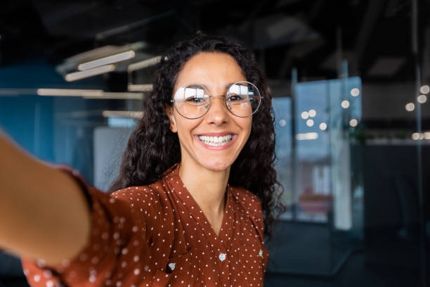istock Business woman in the office talking on a video call, using application on smartphone for online remote communication, looking at the camera and smiling, a Latin American inside wearing glasses 1456423694