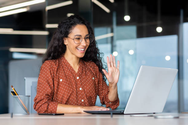 successful and beautiful hispanic woman working inside modern office building, businesswoman using laptop for video call smiling and waving, greeting gesture, online conference with colleagues - zwaaien gebaren stockfoto's en -beelden