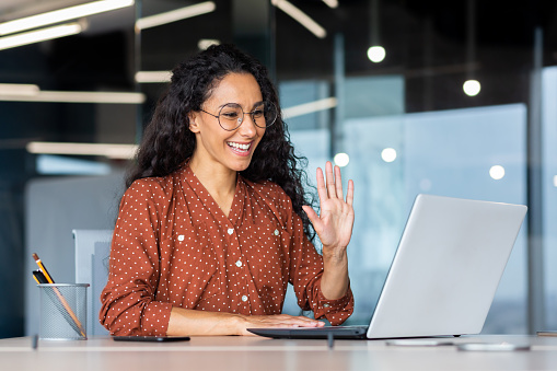 Successful and beautiful hispanic woman working inside modern office building, businesswoman using laptop for video call smiling and waving, greeting gesture, online conference with colleagues