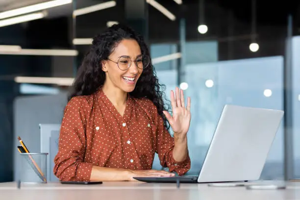 Photo of Successful and beautiful hispanic woman working inside modern office building, businesswoman using laptop for video call smiling and waving, greeting gesture, online conference with colleagues