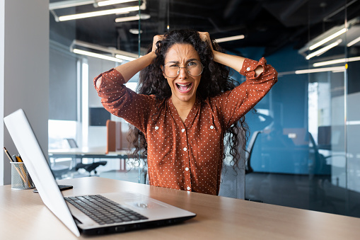 Angry businesswoman yelling at camera, latin american woman holding her head angry working inside modern office using laptop at work.