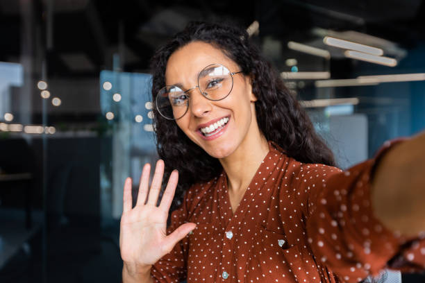 istock Business woman in the office talking on a video call, using application on smartphone for online remote communication, looking at the camera and smiling, a Latin American inside wearing glasses 1456423721