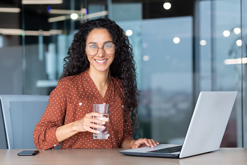 Portrait of Hispanic woman inside modern office at work, businesswoman smiling and looking at camera holding a glass of pure filtered water in her hand.