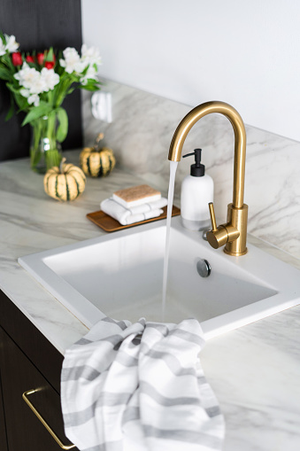 selective focus on golden shiny faucet with water stream at kitchen with modern interior and decor with flowers and pumpkins on background