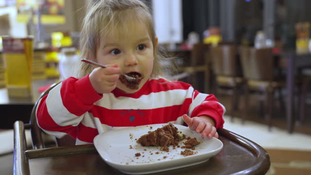 Toddler girl eating chocolate cake, while siting in high chair at cafe