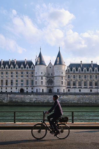 Paris, France - May, 2022: A man riding a bicycle on banks of seine river in front of  La Conciergerie, former prison transformed into courts