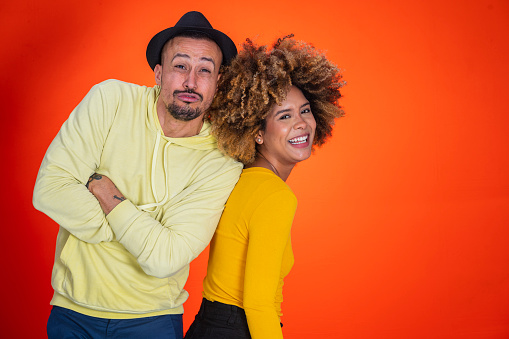 Couple of Latino friends from Bogota Colombia between the ages of 20-39 smiling and enjoying themselves while standing in orange studio background wearing casual clothing