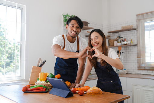 Happy asia young couple cooking together with vegetables in cozy kitchen, vegetarian food eating concept, showing heart shape to camera, true love and valentine concept.