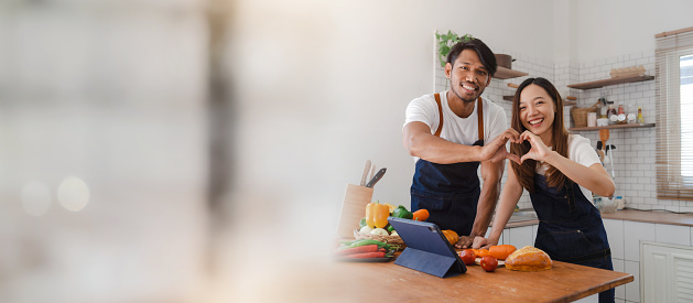 Happy asia young couple cooking together with vegetables in cozy kitchen, vegetarian food eating concept, showing heart shape to camera, true love and valentine concept.