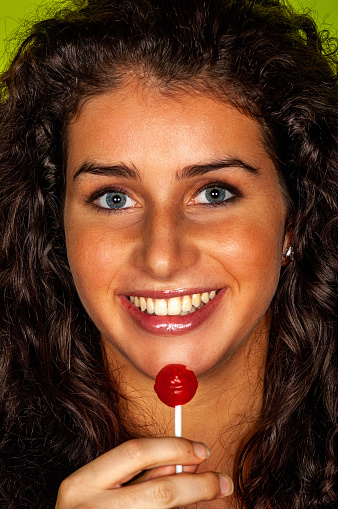 Studio portrait of a beautiful young woman sucking on a red lollipop against a green background