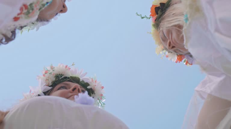 Three happy and smiling women in traditional white shirts and wreaths dance against a blue sky with clouds. Bottom view. Historical reconstruction Slavic Feast of Ivan Kupala