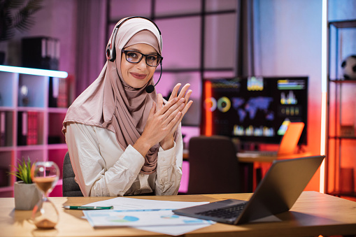 Happy muslim business woman in hijab getting good news, looking at camera in evening office clapping her hands, while celebrating successful completion of project.