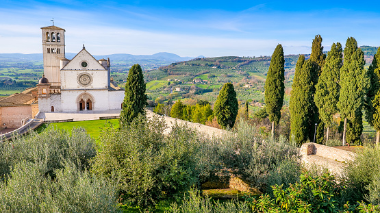 A beautiful and peaceful panorama of the Basilica di San Francesco (Basilica of St Francis) in the medieval town of Assisi, in Umbria, surrounded by olive trees and Italian cypresses. Built in the Italian Gothic style starting from 1228 and completed in 1253, the Basilica, which preserves the mortal remains of the Saint of the Poor from 1230, is composed of the Basilica Inferiore (Lower Basilica) and the Basilica Superiore (Upper Basilica), perfectly integrated. Over the centuries Assisi and the spirituality of its sacred places have become a symbol of peace, a point of reference for tolerance and solidarity between peoples and between the different confessions of the world. The Umbria region, considered the green lung of Italy for its wooded mountains, is characterized by a perfect integration between nature and the presence of man, in a context of environmental sustainability and healthy life. In addition to its immense artistic and historical heritage, Umbria is famous for its food and wine production and for the high quality of the olive oil produced in these lands. Since 2000 the Basilica and other Franciscan sites of Assisi have been declared a World Heritage Site by UNESCO. Wide angle image in 16:9 and high definition format.
