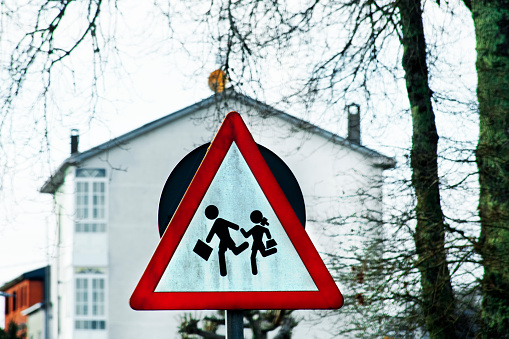 Triangle warning pedestrians road sign, children,school building nearby. Galicia, Spain. Bare tree trunk and branches, building exterior in the background.