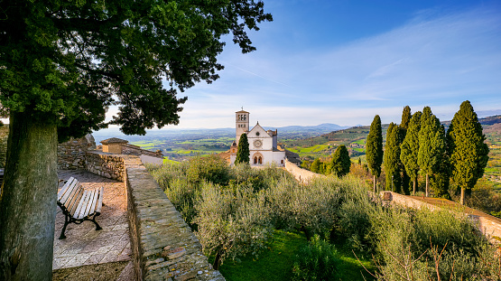 A beautiful and peaceful panorama of the Basilica di San Francesco (Basilica of St Francis) in the medieval town of Assisi, in Umbria, seen from a stone terrace and surrounded by olive trees and Italian cypresses. Built in the Italian Gothic style starting from 1228 and completed in 1253, the Basilica, which preserves the mortal remains of the Saint of the Poor from 1230, is composed of the Basilica Inferiore (Lower Basilica) and the Basilica Superiore (Upper Basilica), perfectly integrated. Over the centuries Assisi and the spirituality of its sacred places have become a symbol of peace, a point of reference for tolerance and solidarity between peoples and between the different confessions of the world. The Umbria region, considered the green lung of Italy for its wooded mountains, is characterized by a perfect integration between nature and the presence of man, in a context of environmental sustainability and healthy life. In addition to its immense artistic and historical heritage, Umbria is famous for its food and wine production and for the high quality of the olive oil produced in these lands. Since 2000 the Basilica and other Franciscan sites of Assisi have been declared a World Heritage Site by UNESCO. Super wide angle image in 16:9 and high definition format.