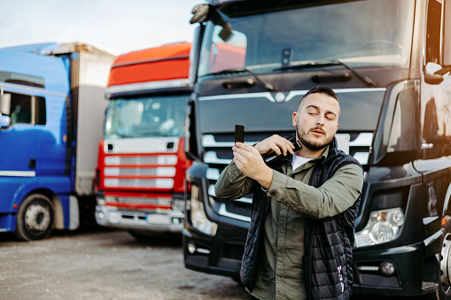 Young truck driver prepares for his next tour. He stands in front of the truck and shaving his face with electric shaver.