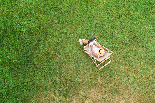 Woman in a white bikini in hat with laptop sitting on deck chair on the green grass sunbathes at summer day. Top view, drone, aerial view.