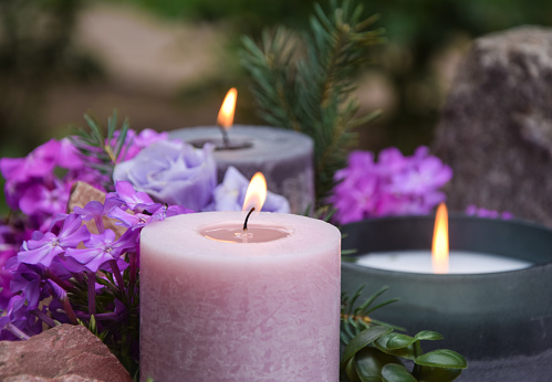 Closeup burning candles purple flowers stone on blurred background. Focus on light candlewick. Beautiful composition with violet candles for spa treatment. Zen relax memorial concept.