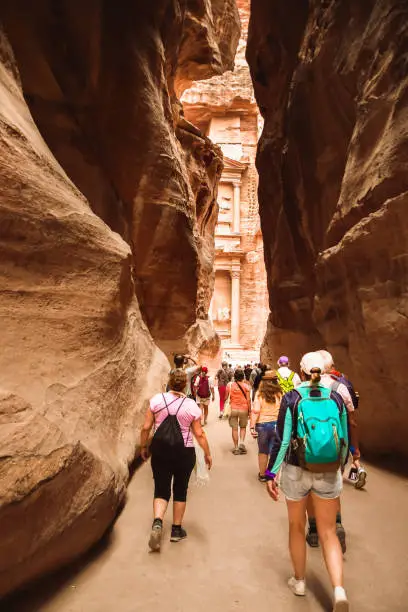 Tourists walk towards treasury landmark in Petra through The Siq, the narrow slot-canyon that serves as the entrance passage to the hidden city of Petra, Jordan