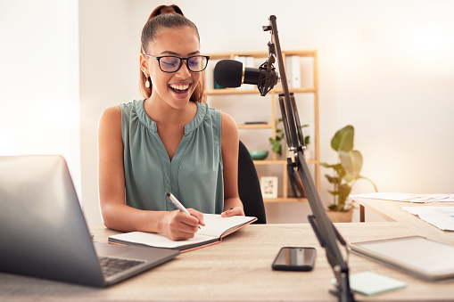Podcast microphone, laptop and woman writing in notebook for ideas, research or information. Influencer, presenter or journalist planning blog for live streaming, radio talk show or online broadcast.