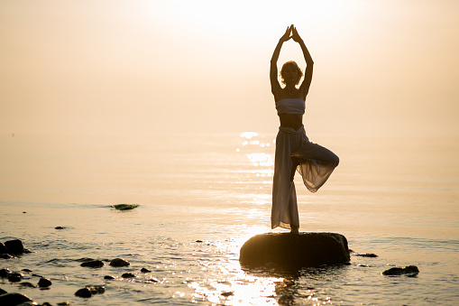 Silhouette of woman who practices yoga tree pose on the beach standing on stone at sunset. Healthy lifestyle concept.