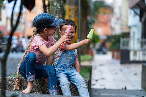 Two Latin brothers from Bogotá Colombia, between 7 and 3 years old, enjoy eating their popsicle while playing in the street wearing comfortable clothes