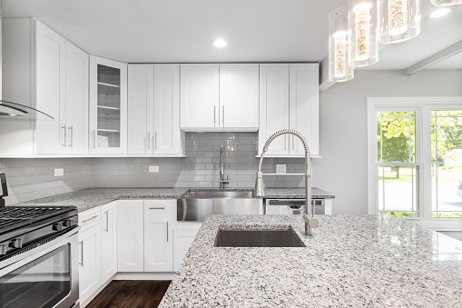 An elegant, modern kitchen featuring grey and white speckled granite, stainless steel appliances, white cabinets and stainless steel appliances with a fancy light.
