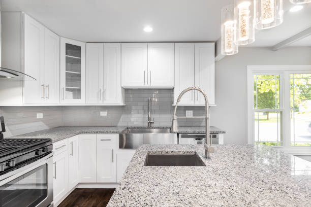 A white kitchen with a stainless steel apron sink, granite countertops and white cabinets. An elegant, modern kitchen featuring grey and white speckled granite, stainless steel appliances, white cabinets and stainless steel appliances with a fancy light. granite rock stock pictures, royalty-free photos & images