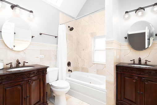 A luxury bathroom with two mahogany wood cabinets and faucets, circular mirrors, and tiled walls and floor.