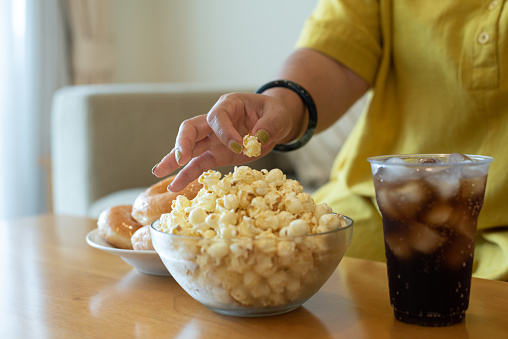 Indoors, close-up shot with copy space of unrecognizable woman picking popcorn in living room. Unhealthy lifestyle, bad habits, diet, junk food, couch potato, health problem concept.