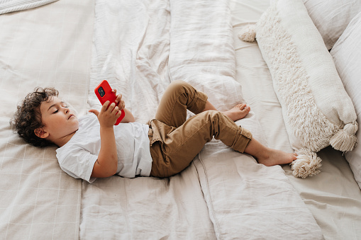 Bored curly little boy laying on bed holds phone makes video call, wants to go outside to play with friends. Isolation, illness. Handsome Italian kid playing games on smartphone. Children and gadgets.