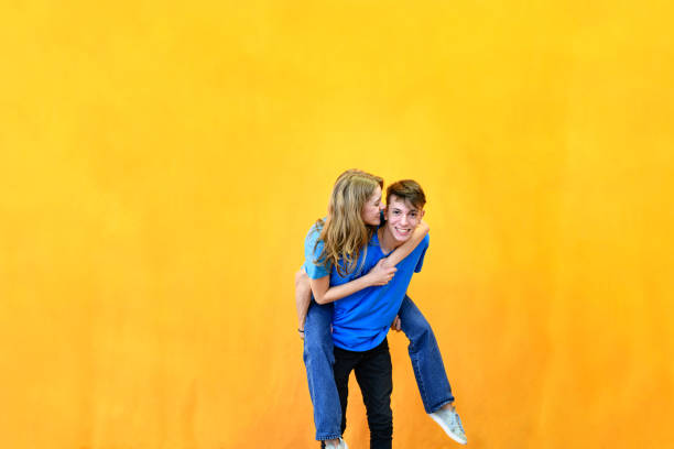 Couple of young lovers, piggyback, having fun. Studio photo. Blue outfit and orange background.