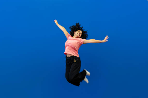 Young curly hair woman jumping high in joy, having a wonderful time, excited. Studio photo. Pink outfit and blue background.