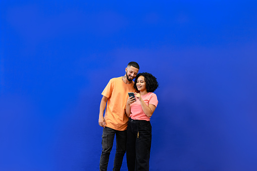 Young couple having fun looking at the mobile. Studio photo. Orange clothing and blue background.