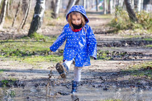 Little caucasian girl playing with the puddles created by the rain.  Girl in a raincoat and rain boots.  Children's games in the park.