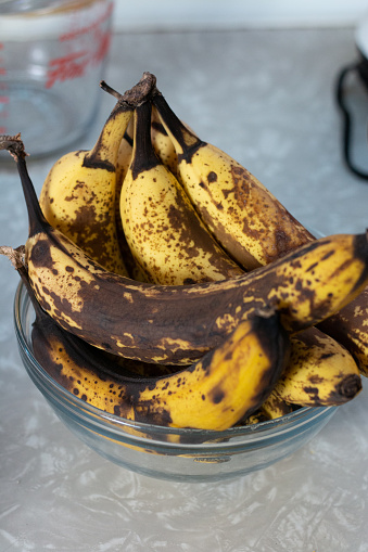 a bunch of yellow-brown bananas are sitting in a glass bowl on a marbled countertop; there is a glass measuring cup behind it, and a visible black electrical cord in the right corner.