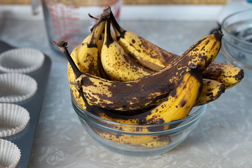 a bunch of yellow-brown bananas are sitting in a glass bowl on a marbled countertop; there is a stack of glass bowls to the right, a glass measuring cup behind it, and a lined cupcake tray to the left.