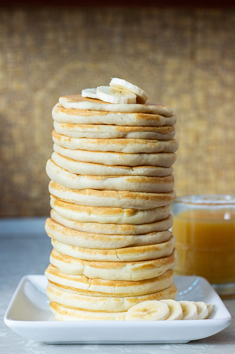 a tall stack of pancakes sit on a white square plate. a few slices of banana sit on top of the stack and around the front right side of the plate. a short glass of orange juice sits behind and to the right of the stack.