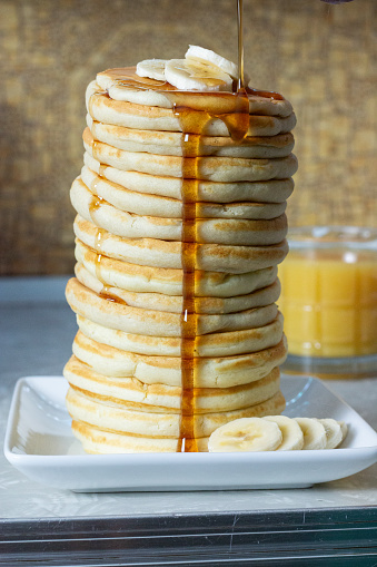 a tall stack of pancakes sit on a white square plate. a few slices of banana sit on top of the stack and around the front right side of the plate. a short glass of orange juice sits behind and to the right of the stack. syrup is being poured over the pancakes and it is dripping down the front of the stack.
