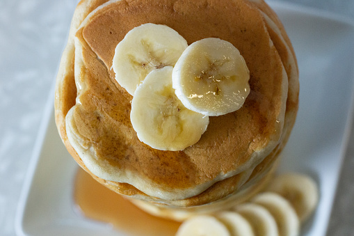 a close up of a stack of pancakes, being shown from overhead. they sit on a white, square plate, and there are three circular slices of banana sitting on top of the stack, more banana slices sit around the stack on the bottom right and some syrup has pooled to the left.