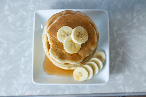 a stack of pancakes is being shown from overhead. they sit on a white, square plate, and there are three circular slices of banana sitting on top of the stack, more banana slices sit around the stack on the bottom right and some syrup has pooled to the left.
