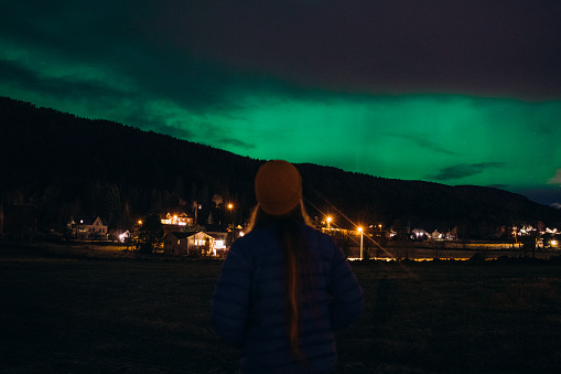 Female in hat admiring majestic bright colorful Northern Lights during wintertime above the mountain village in Scandinavia