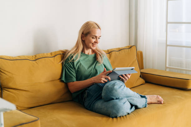istock Attractive middle-aged caucasian woman sitting on a sofa works on a computer tablet. 1456588080