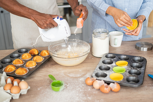Close shot of young multiracial couple cooking together and having good time in the kitchen. Man mixing eggs in a bowl and adding flour, while his girlfriend  helping him with the muffin's  tin