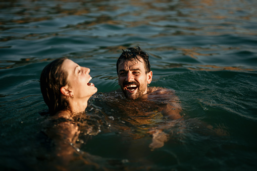 Romantic side shot of white husband and wife. They are smiling while swimming in the sea. Sweet sunset shot