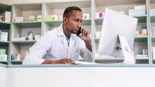 istock Multiracial male pharmacist, checking medicine on a computer and talking on the phone at a pharmacy. Multiracial pharmacist working in drugstore at a pharmacy. 1456591961