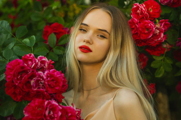 A Russian model checking out red flowers in a community garden. She is wearing medium length blond hair, makeup, and a white short sleeved dress.