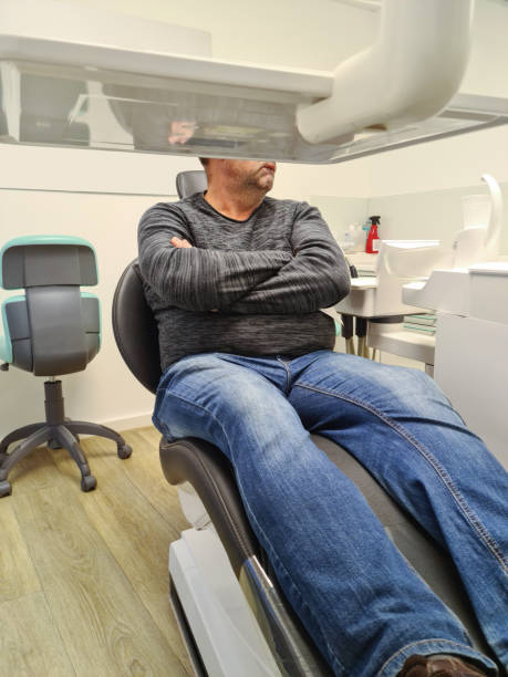 The patient is sitting in the dentist's chair, and from his posture one can understand that he is afraid. His arms are crossed over his chest, his lips are tightly compressed, he unconsciously bit his lower lip. stock photo